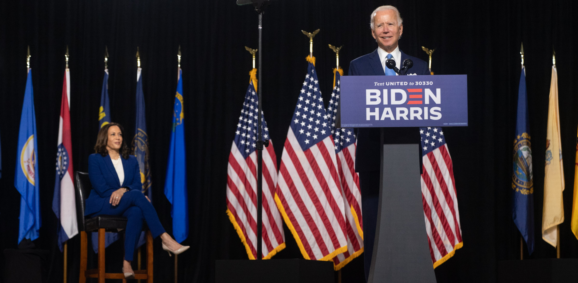 Joe Biden standing at a podium with Biden Harris logo on the front. There are a series of flags behind him.